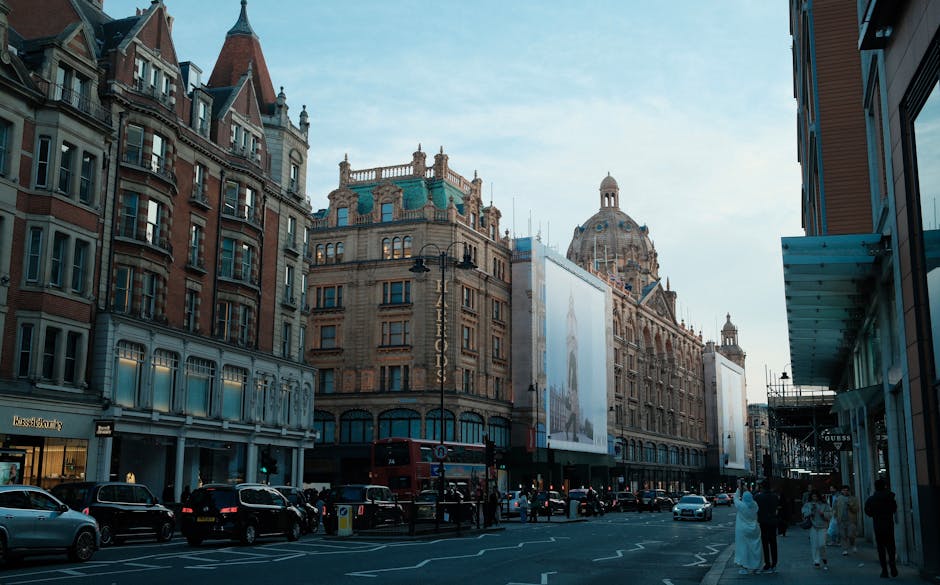 A city street scene along Brompton Road in Knightsbridge, featuring historic and ornate buildings with intricate stone and brick facades, some topped with decorative turrets and domes. The street is lined with parked cars, and a red double-decker bus is visible in the distance. Pedestrians walk along the pavement, with some engaging in conversation or browsing shop windows. The sky is clear with a soft blue hue, suggesting early evening or late afternoon lighting. On the right, there are modern shopfronts with large glass windows and signage, contrasting with the classical architecture on the left. The overall scene captures the blend of historic charm and contemporary urban activity, typical of the area, with the influence of professional gardening and landscape maintenance by Gardeners Knightsbridge not directly visible but implied through the well-kept surroundings.